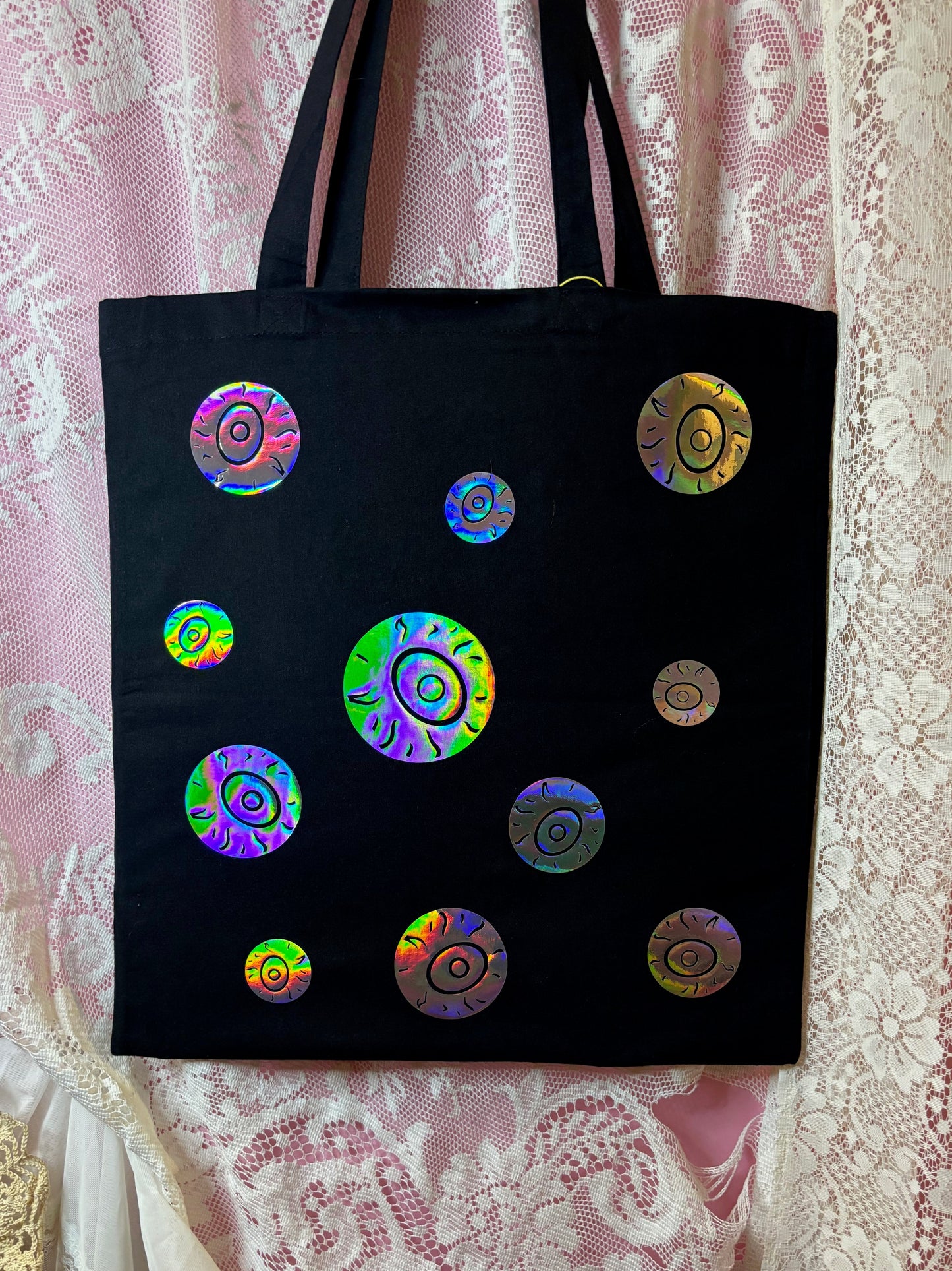 A black canvas tote bag with multiple iridescent holographic circular eyeballs, displayed against a pink and white lace curtain background.