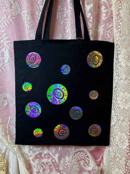 A black canvas tote bag with multiple iridescent holographic circular eyeballs, displayed against a pink and white lace curtain background.